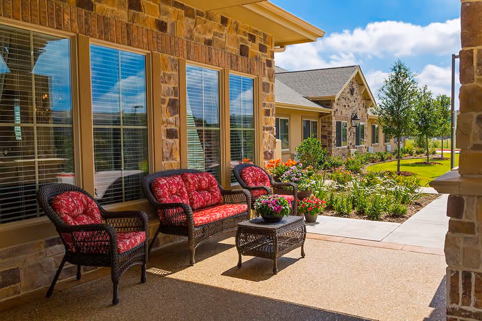 Outdoor patio area at Riverside Oxford Memory Care featuring wicker furniture with red cushions, a small table with potted flowers, stone building exterior, large windows with blinds, and a landscaped garden with flowers and trees under a blue sky with clouds.
