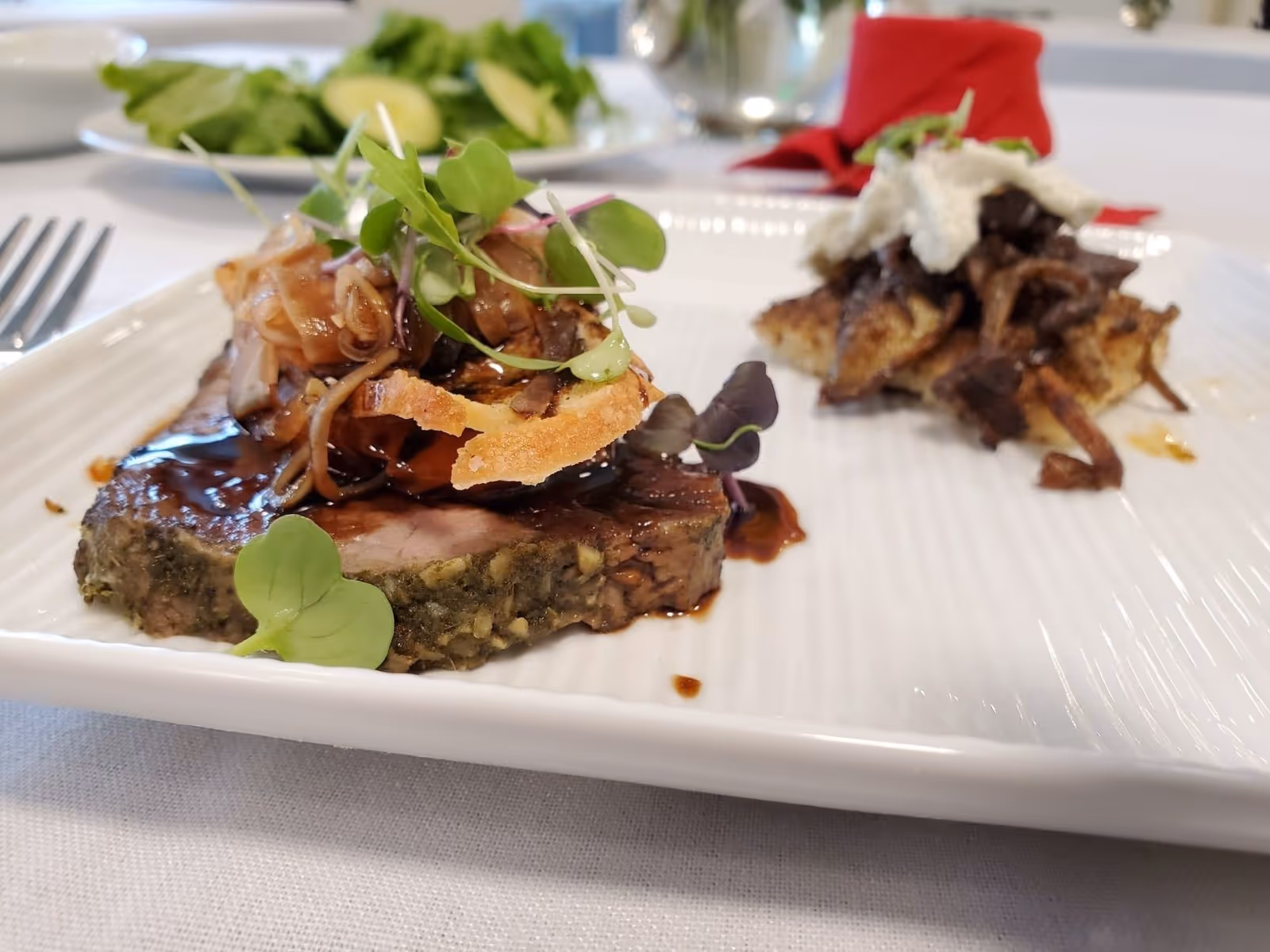 Close-up of a plated gourmet meat dish topped with microgreens and sauce on a white plate at a dining table.