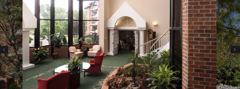 Bright interior lounge with seating, large windows, indoor plants, and a staircase leading to an upper area.