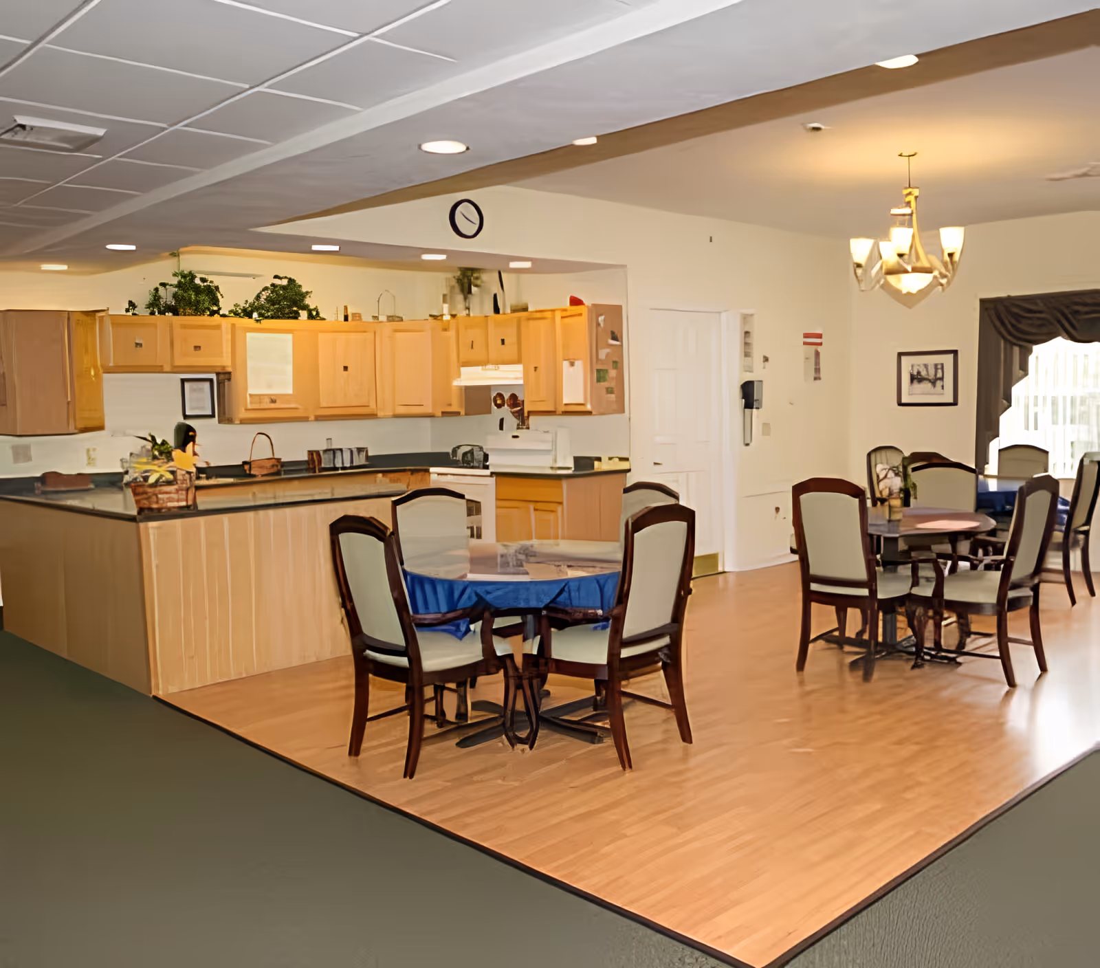 Interior view of a senior living facility dining area and kitchen at The Terrace at Woodland, featuring wooden cabinets, a kitchen counter, several round tables with chairs, a chandelier, and a window with curtains allowing natural light.