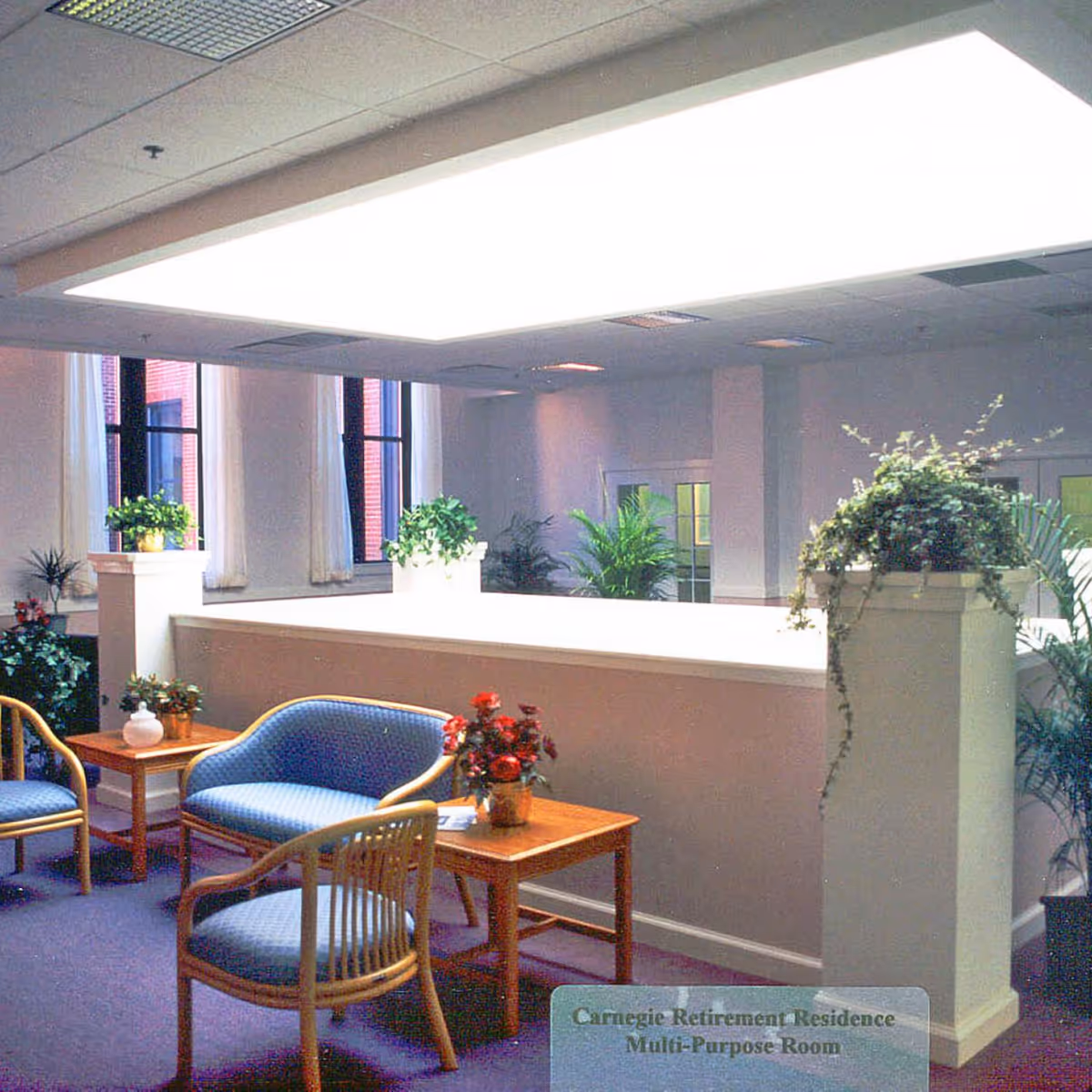 Well-lit senior living common room with seating, tables, and potted plants under a large rectangular ceiling light.