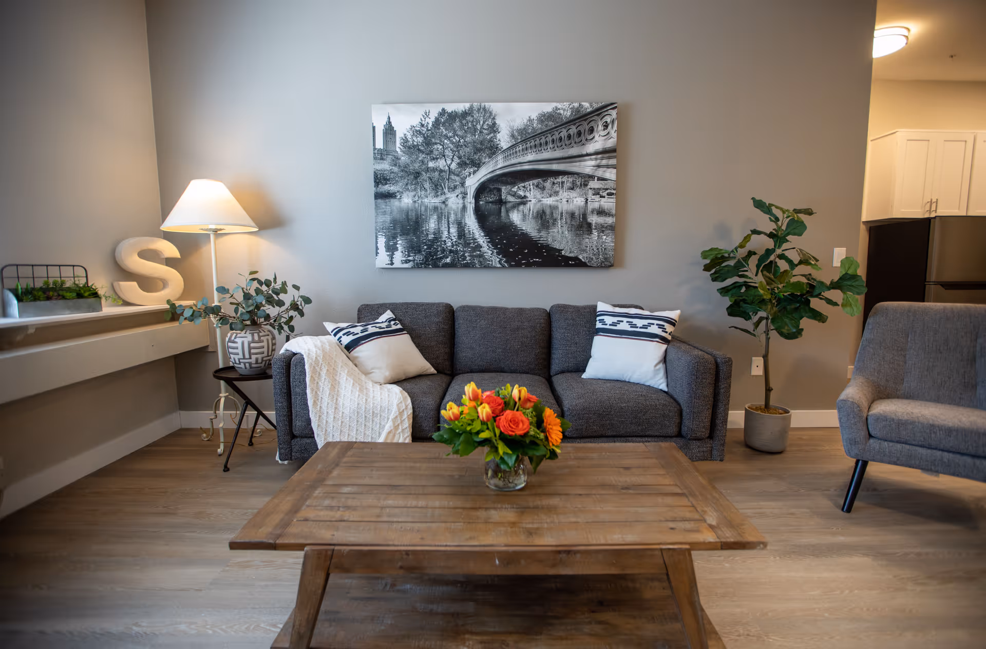 Cozy living room with a gray sofa and pillows, wooden coffee table with a vase of flowers, armchair, lamp, and wall art.