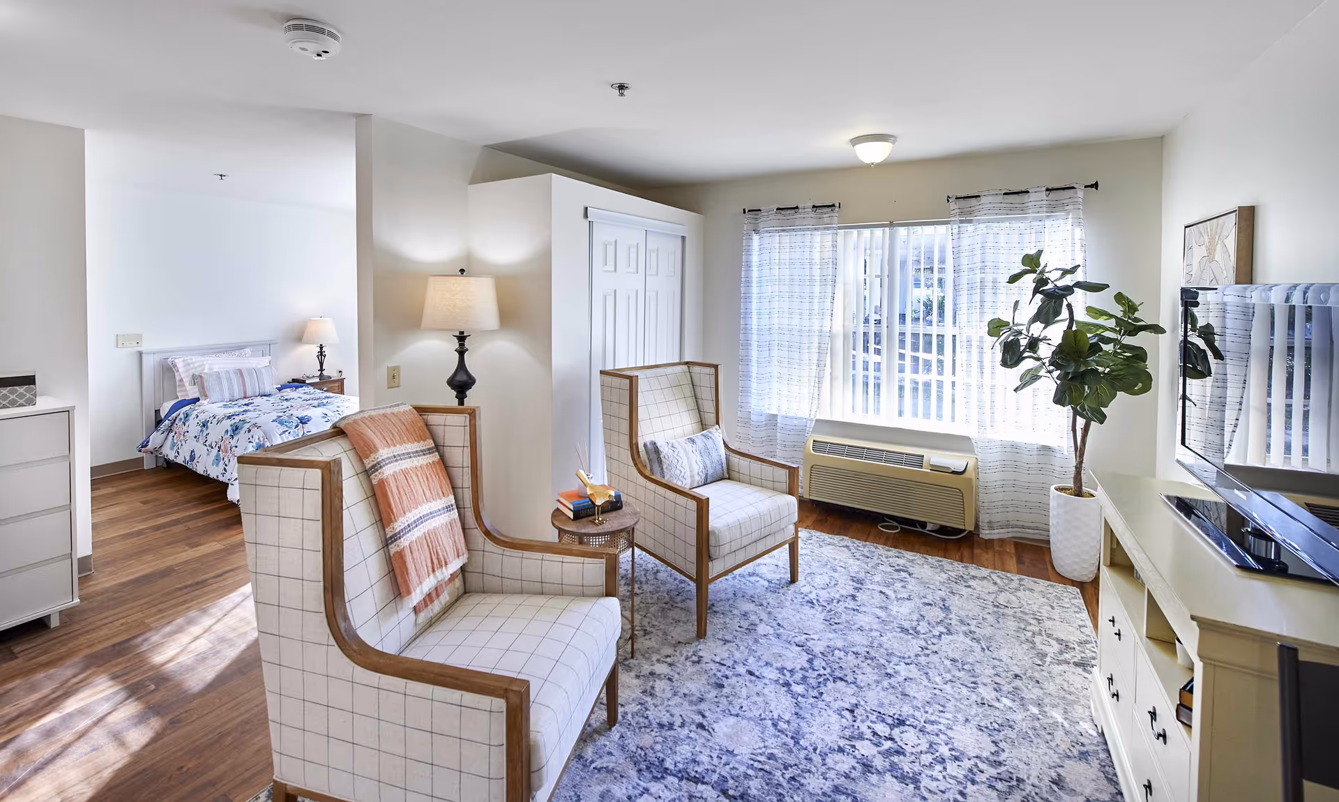 A bright and cozy living area with two cushioned armchairs, a small round side table, a large window with sheer curtains, a potted plant, and a TV on a white cabinet. In the background, there is a bedroom with a bed, nightstand, and lamp. The room has wooden flooring and light-colored walls.