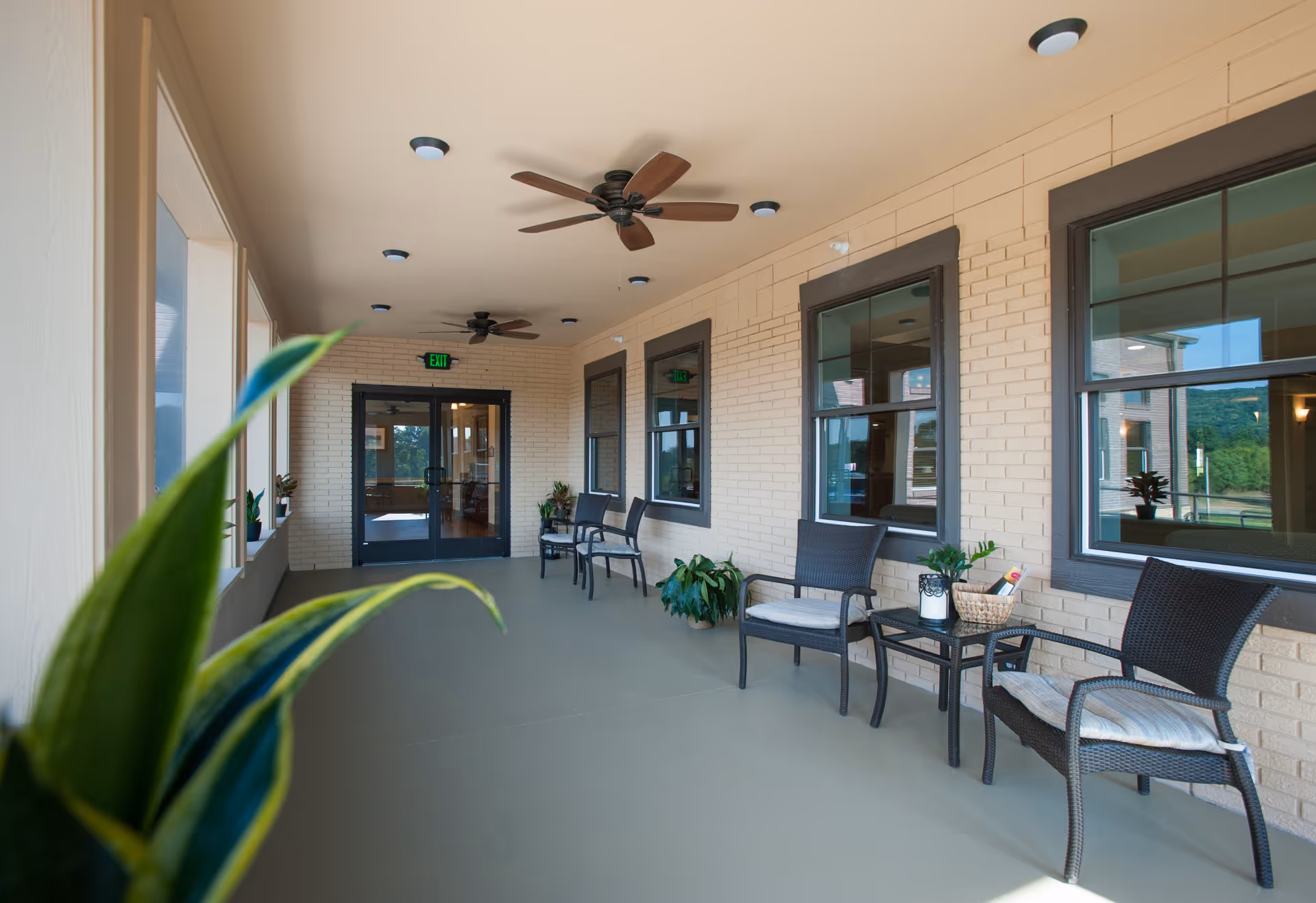 Covered outdoor patio area with ceiling fans, several chairs with cushions, small tables, potted plants, and large windows looking into the building.