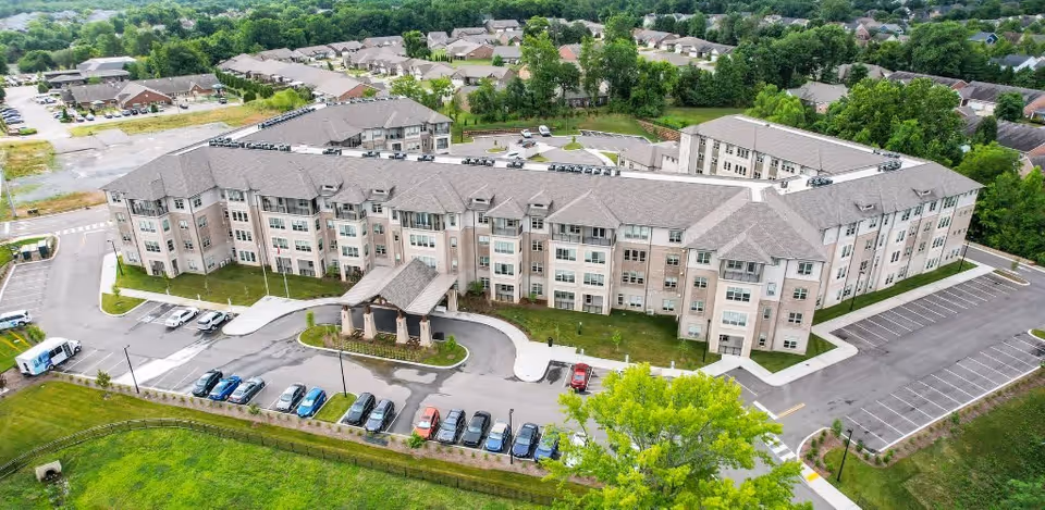 Aerial view of a large, multi-story senior living facility building with a covered entrance and surrounding parking lots. The building is surrounded by green lawns and trees, with a residential neighborhood visible in the background.