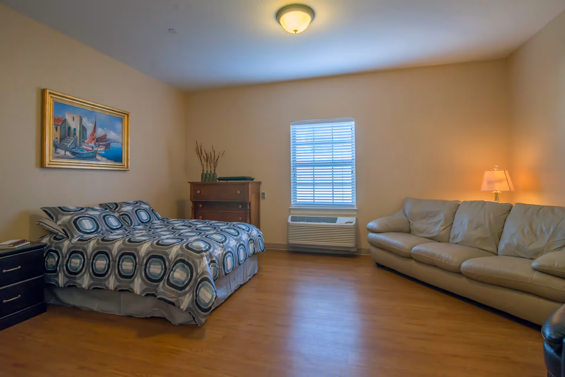 A bedroom with a bed covered in a patterned blue and gray bedspread, a wooden dresser with decorative vases, a window with blinds, a beige leather couch, a side table with a lamp, and a framed painting of boats on the wall.