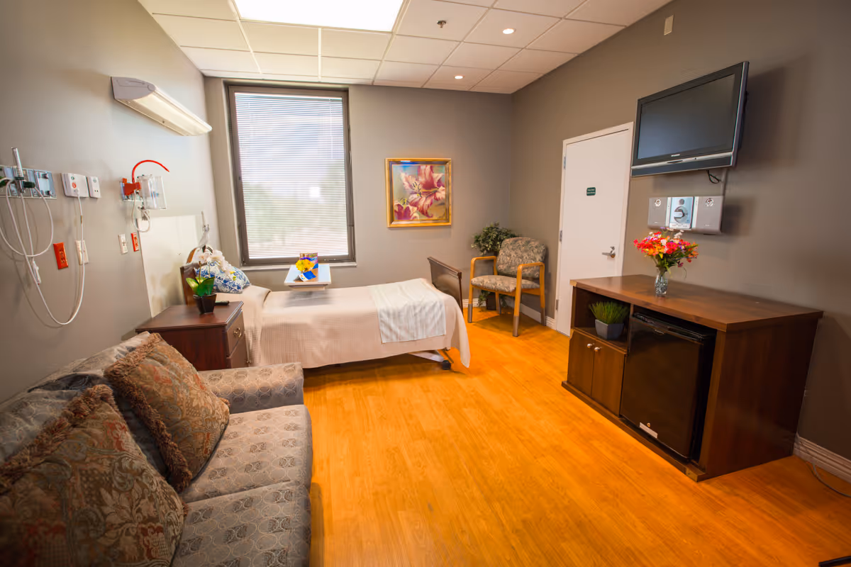 A well-lit patient room with a hospital bed near a window, a wooden nightstand with a plant and flowers, a patterned sofa with cushions, a wooden cabinet with a mini fridge and a vase of flowers, a wall-mounted TV, and a chair next to a closed door. The room has wood flooring and gray walls with a framed floral painting.