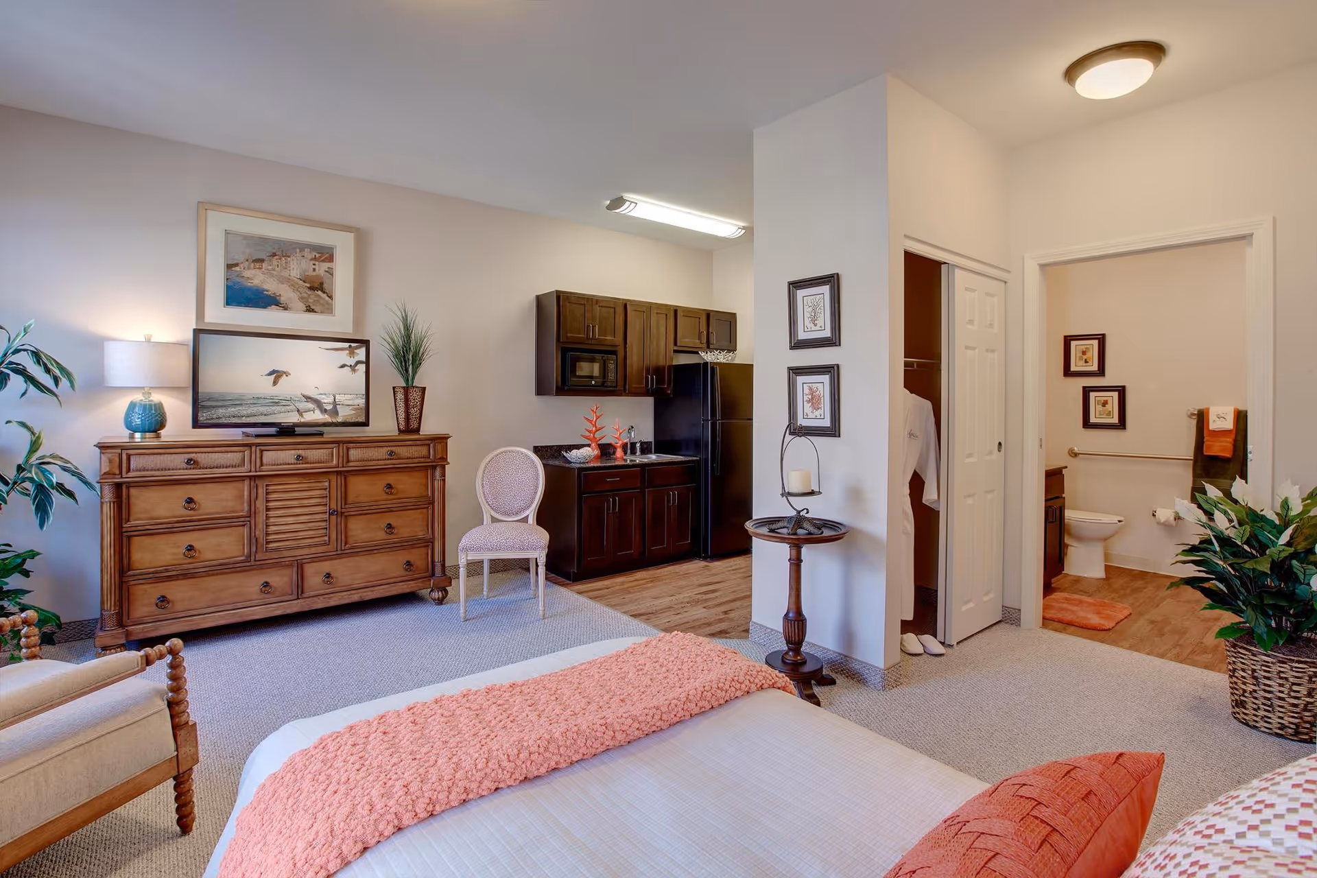 A senior living facility room featuring a bed with coral-colored bedding, a wooden dresser with a lamp and framed pictures above it, a small kitchenette with dark cabinets and a black refrigerator, a chair, a small round table with a candle, and an open doorway leading to a bathroom with a toilet and framed pictures on the wall. There are also plants placed around the room.