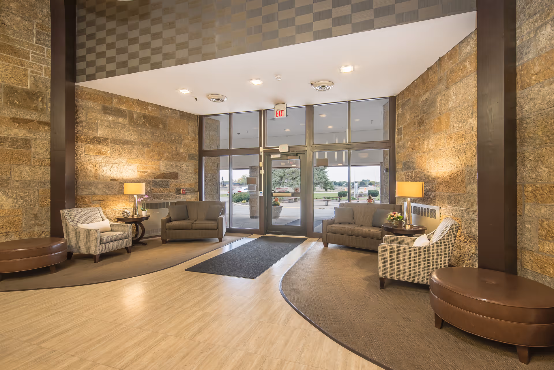 A spacious and well-lit lobby area with stone walls, two gray sofas, two patterned armchairs, two round side tables with lamps and flowers, and large glass double doors leading outside.