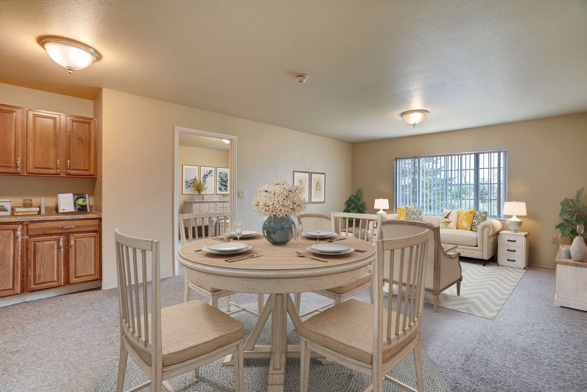 A bright and cozy senior living space featuring a round dining table set with four chairs and place settings. The room includes a living area with a beige sofa adorned with yellow and patterned cushions, two side tables with lamps, a large window with vertical blinds, and decorative plants. Wooden cabinets and a countertop are visible to the left, and a doorway leads to another room with framed artwork on the wall.
