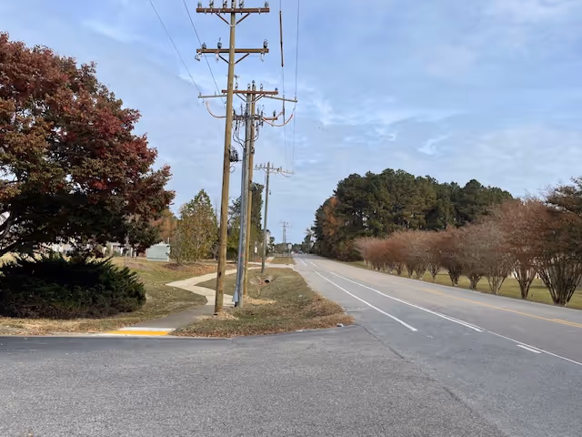 A quiet road with utility poles and a sidewalk bordered by trees and grass under a cloudy sky.