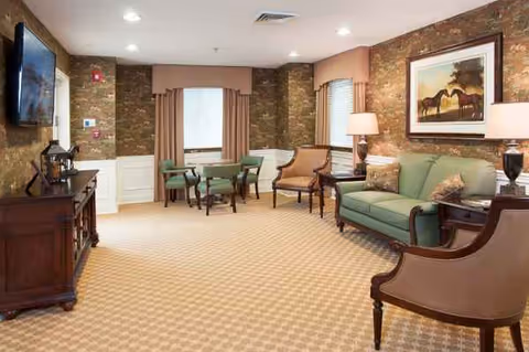 Senior living common room with green sofas, chairs, lamps, brick walls, and artwork.