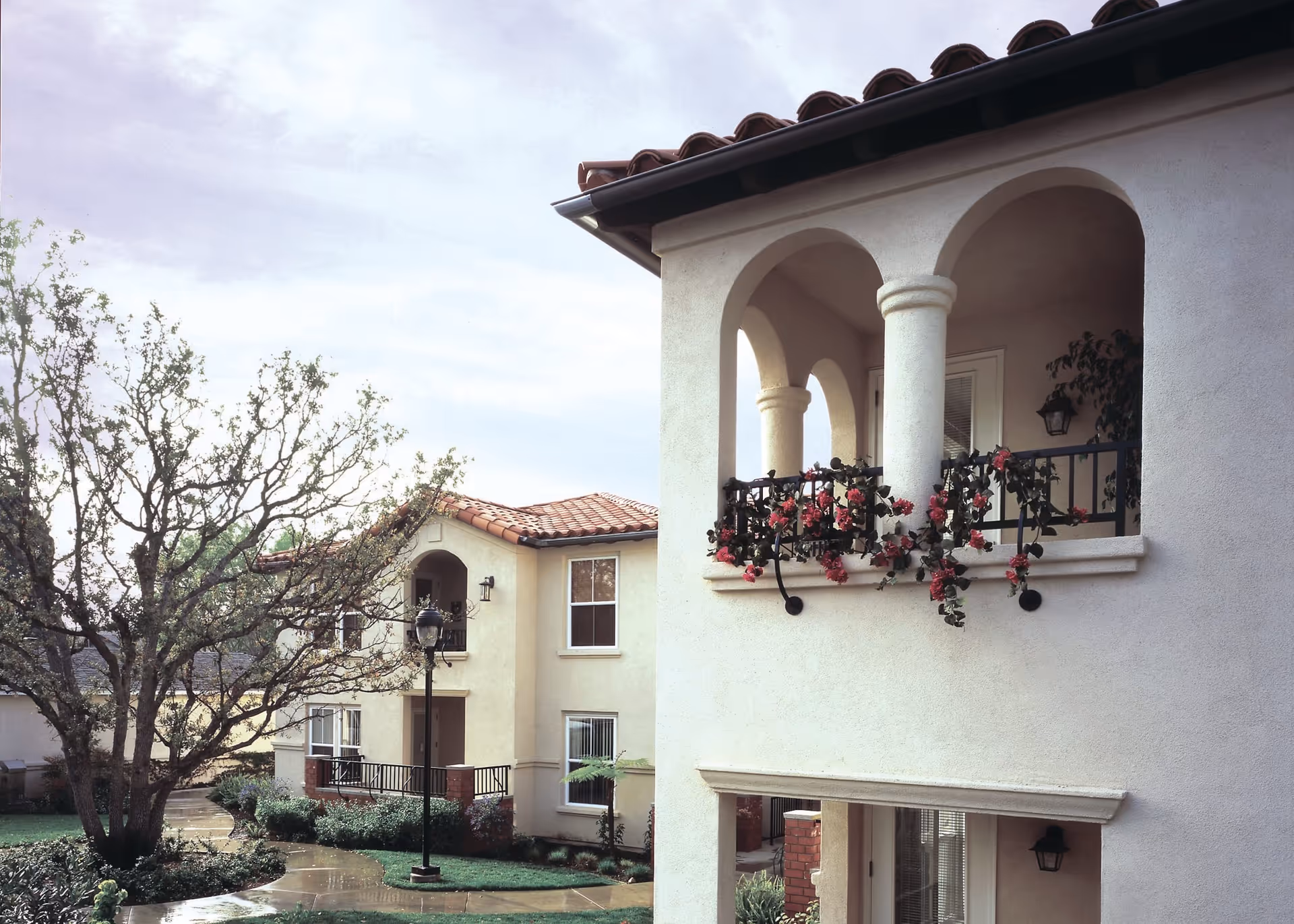 Exterior view of a retirement community building with beige stucco walls and red tile roof. The building features arched balconies decorated with pink flowers. A tree and a lamppost are visible along a curved walkway surrounded by greenery.