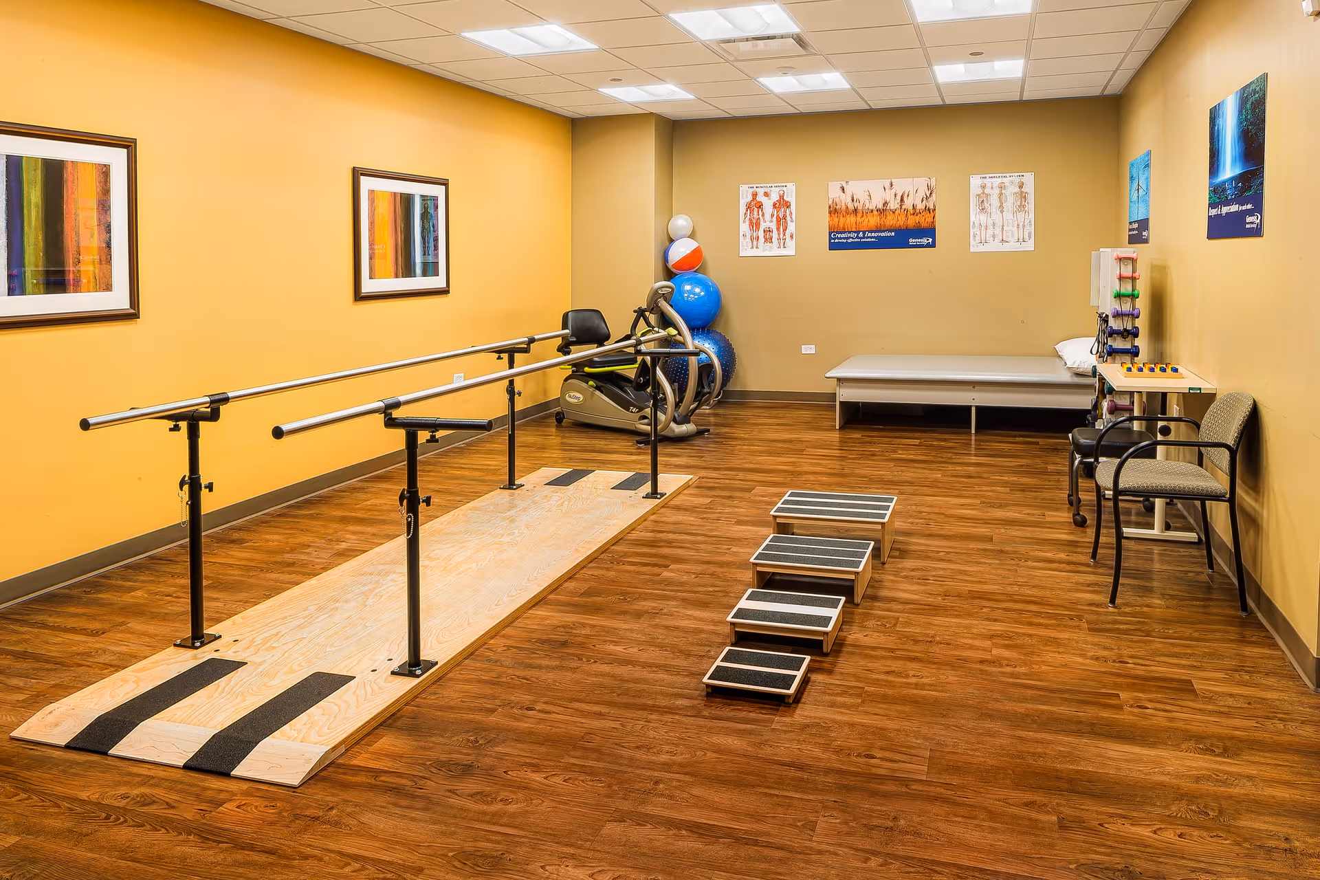 A physical therapy or rehabilitation room with wooden flooring, parallel bars for walking exercises, a set of four graduated steps, exercise balls, a stationary bike, a treatment table, chairs, and anatomical posters on the walls.