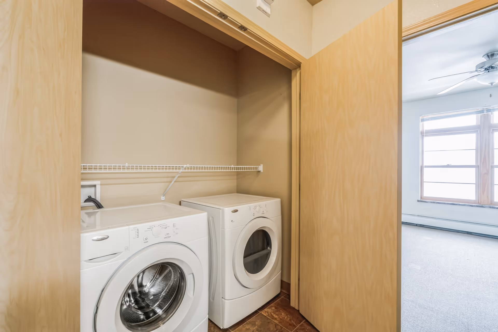 Laundry area with a front-loading washing machine and dryer inside a closet with wooden sliding doors. Adjacent room with carpeted floor, large window, and ceiling fan is visible.