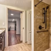 View from a bathroom showing a walk-in shower with brown tiles and a grab bar, looking out into a hallway with wooden flooring and a partially open door leading to another room.