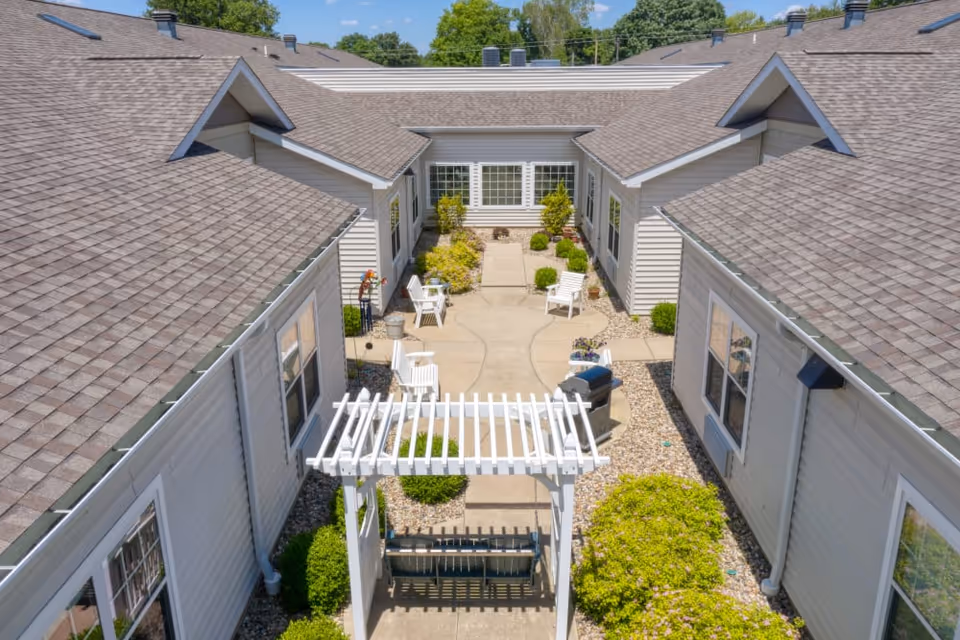 A peaceful outdoor courtyard area at Addington Place of Edwardsville featuring a paved walkway, white chairs, a grill, and well-maintained bushes and plants, surrounded by single-story buildings with gray roofs and beige siding under a clear blue sky.