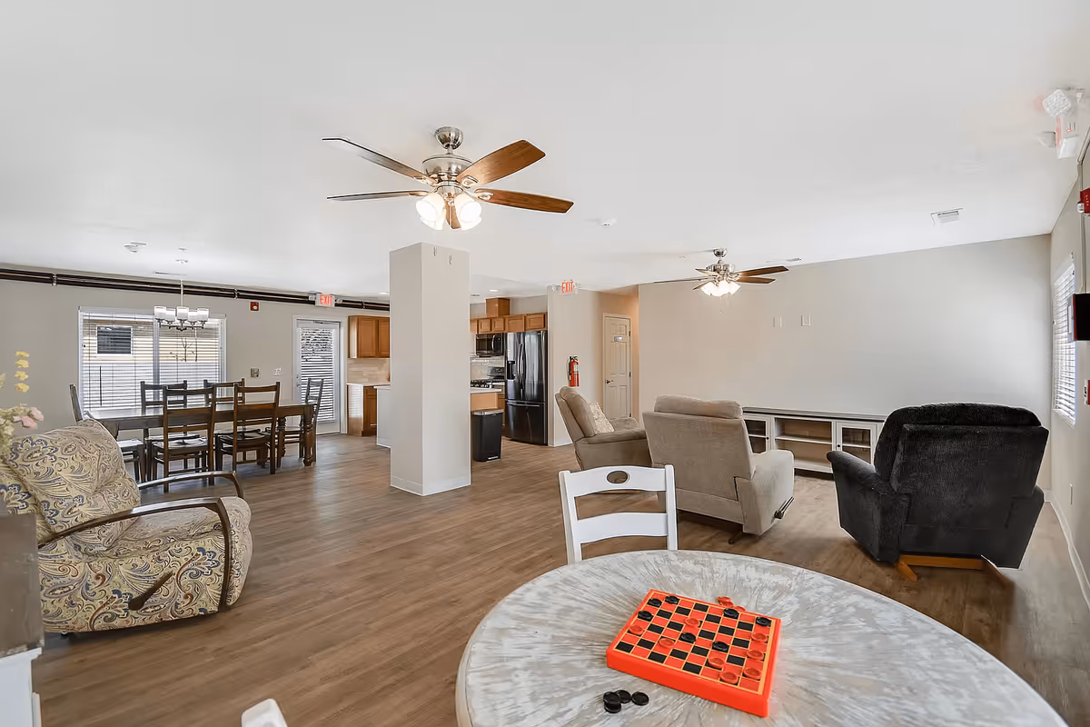 A spacious common area in a senior living facility featuring a round table with a checkers game in the foreground, several armchairs and sofas arranged around a TV stand, a dining table with chairs near windows, ceiling fans with lights, and a kitchen area with wooden cabinets and black appliances in the background.