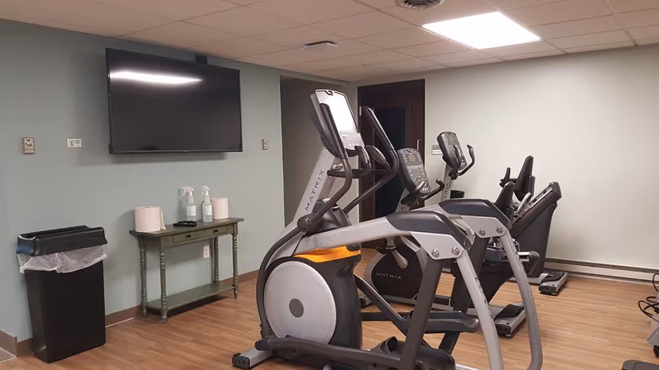 A small exercise room with several pieces of cardio equipment including elliptical machines and stationary bikes. There is a wall-mounted flat screen TV above a small green table holding cleaning supplies and paper towels. A black trash can is positioned against the wall on the left side. The room has light green walls and wood-style flooring.