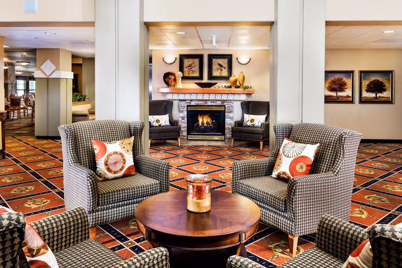 A cozy living room area in a senior living facility with four patterned armchairs arranged around a round wooden coffee table with a candle on it. In the background, there is a lit fireplace with decorative vases and framed bird artwork above it. The room has a red and orange patterned carpet and warm lighting, with additional seating and framed tree artwork visible on the walls.