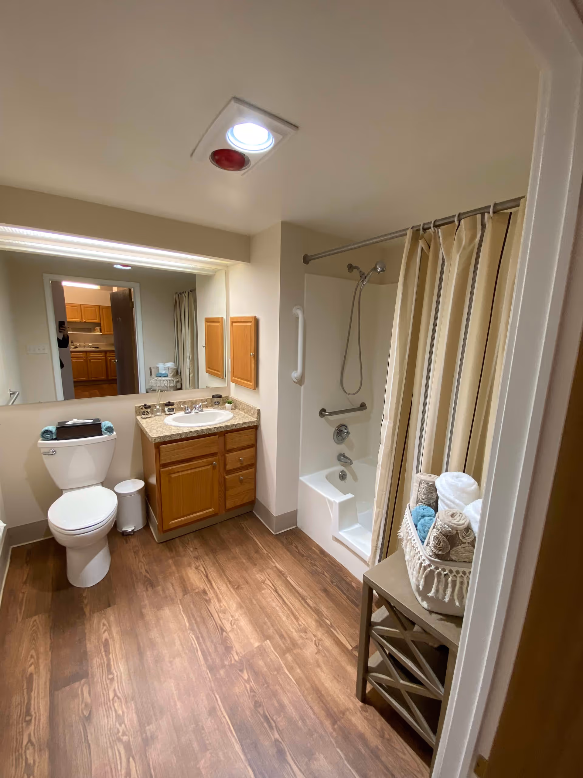 A clean and well-lit bathroom with wood-style flooring. The bathroom features a white toilet with a tissue box and rolled towels on top, a wooden vanity with a sink and countertop, a large mirror above the sink, and a bathtub with a showerhead and beige shower curtain. There is a small table holding a basket filled with rolled towels next to the bathtub. The ceiling has a recessed light and a red emergency light.