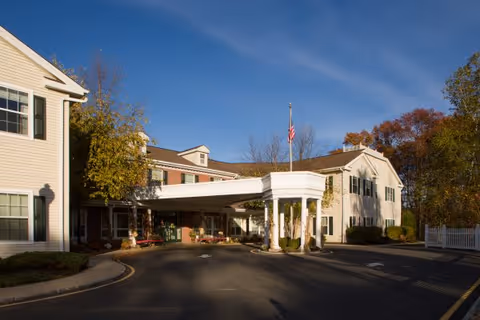 Exterior view of a senior living facility building with a covered entrance and an American flag on a flagpole. The building is two stories tall with beige siding and green shutters. Trees with autumn foliage surround the area under a clear blue sky.