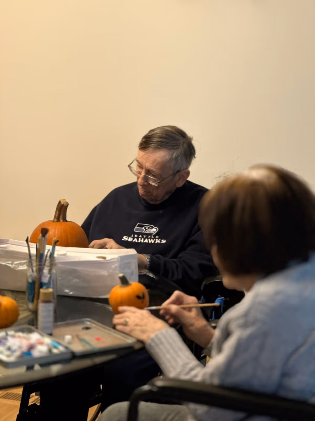 Two elderly individuals seated at a table engaged in painting small pumpkins. One person is wearing a Seattle Seahawks sweatshirt and glasses, while the other is holding a paintbrush and painting a pumpkin. Various painting supplies are visible on the table.