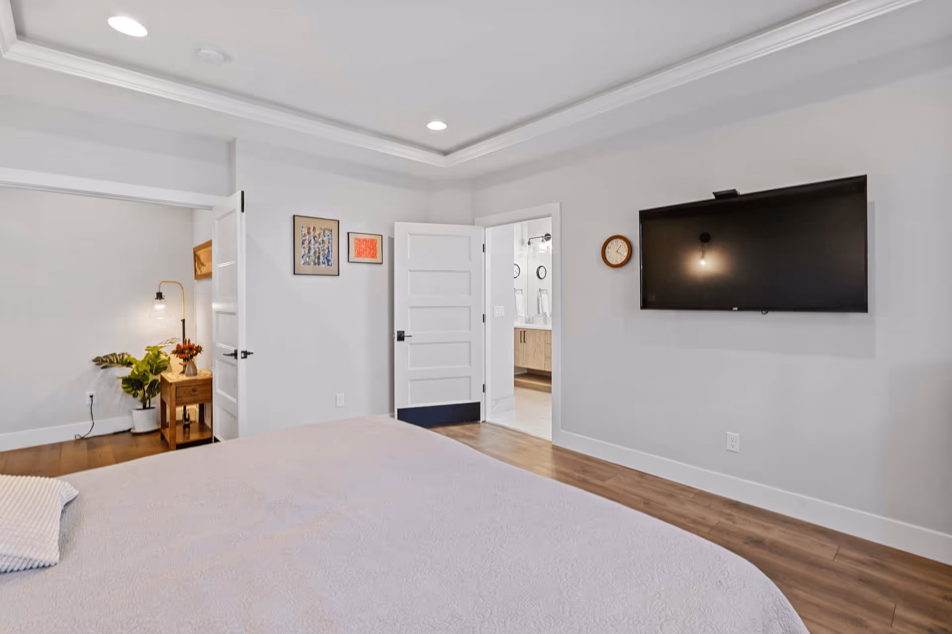 A modern bedroom with a large bed covered in a light-colored quilt. The room has light gray walls, wooden flooring, and recessed ceiling lights. There is a flat-screen TV mounted on the wall opposite the bed, a round wall clock beside it, and two open doors showing a glimpse of an adjacent room with a plant and a nightstand with a lamp and flowers.