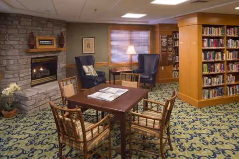 A cozy senior living lounge with a stone fireplace, table, chairs, lamp, and bookshelves.