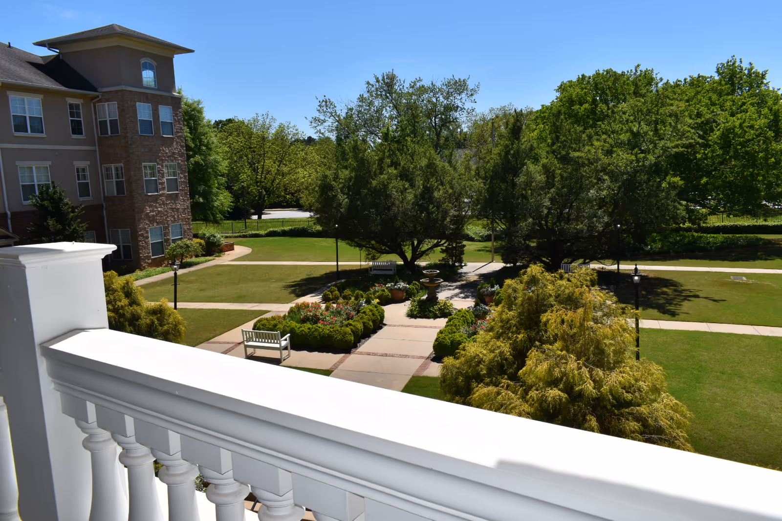 View from a white balcony over a landscaped courtyard with benches, trees, and a multi-story building at left.
