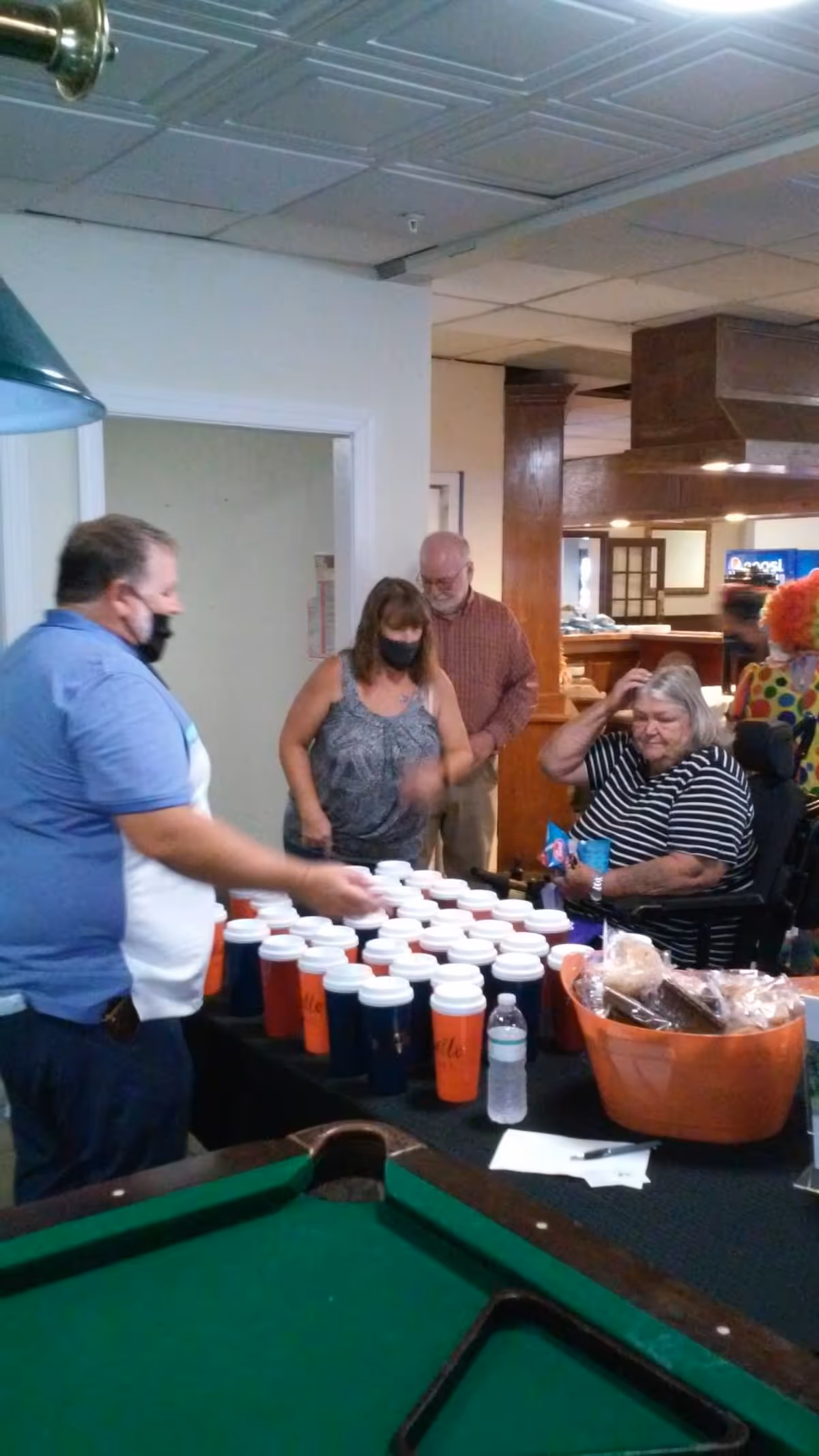 Several people gathered around a table with many cups and a basket of snacks in an indoor common area with a pool table in the foreground. Some individuals are wearing masks.