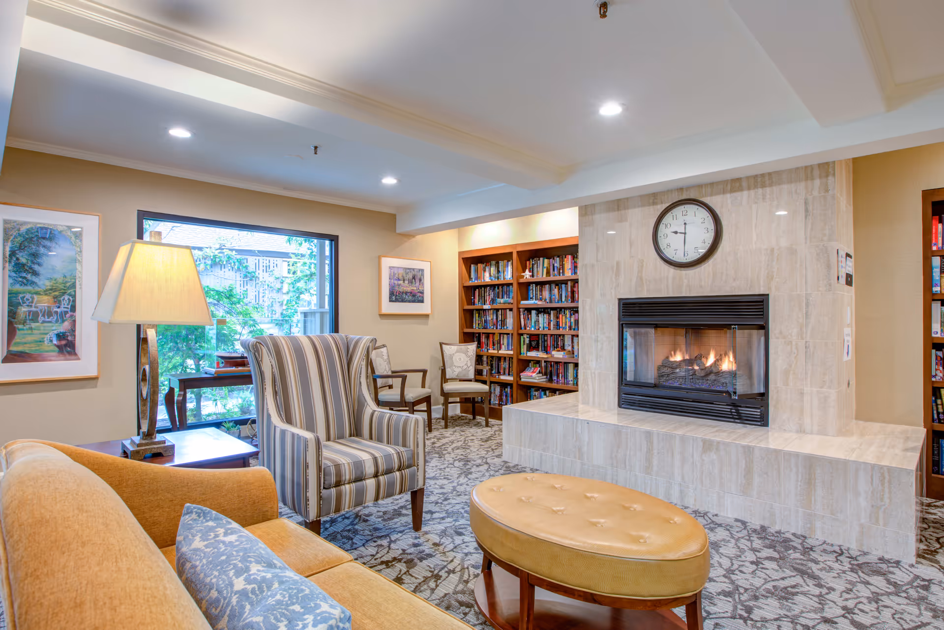 A cozy living room area with a lit fireplace surrounded by beige marble tiles. Above the fireplace is a round clock showing 12:15. In front of the fireplace is a tan oval ottoman. To the left, there is a striped armchair and a mustard-colored sofa with a blue patterned pillow. A table lamp with a beige shade sits on a side table next to the sofa. Behind the armchair, there is a large window showing greenery outside and framed artwork on the walls. Bookshelves filled with books are visible on either side of the fireplace.