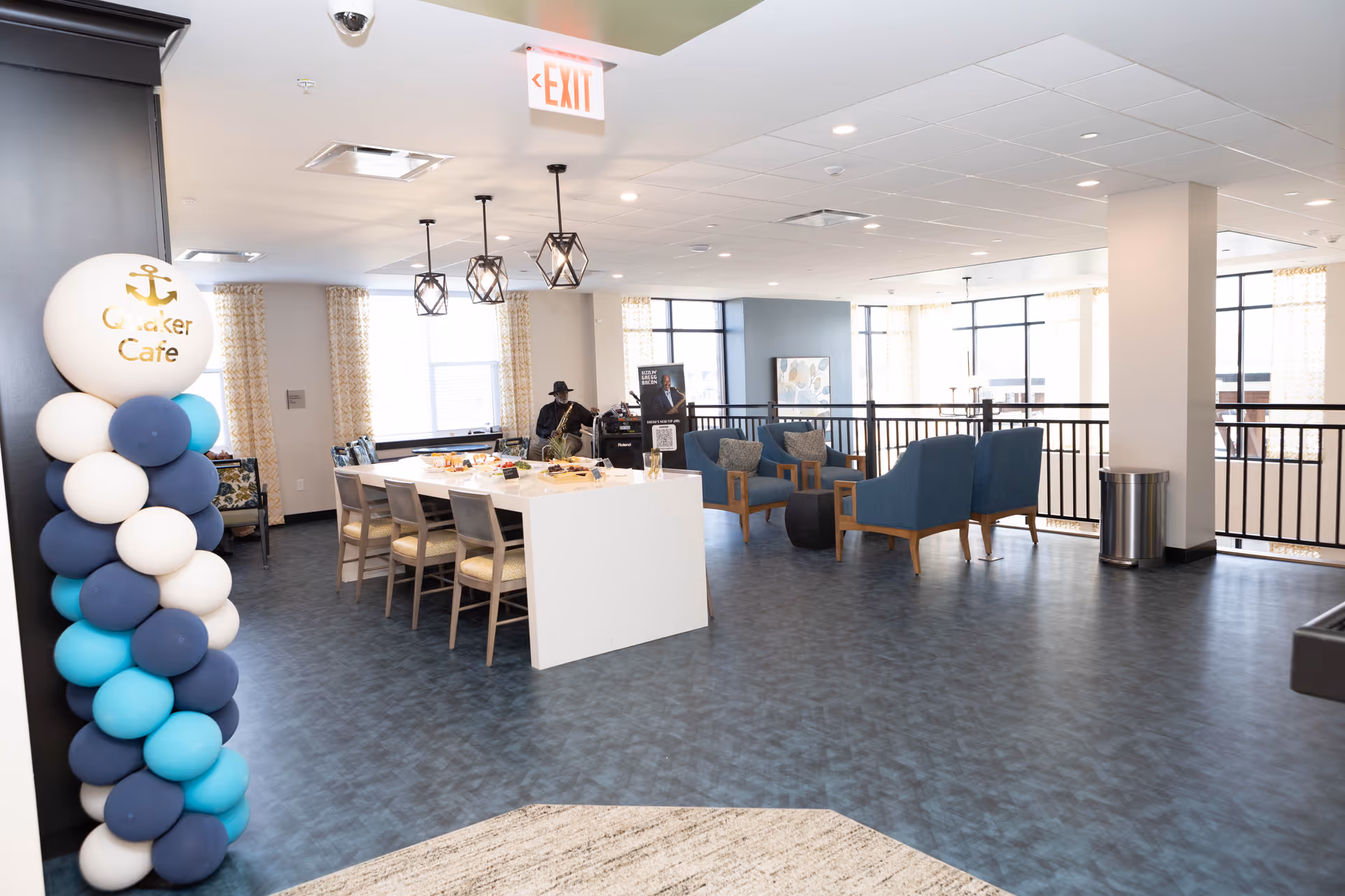 Interior view of a senior living facility lounge area with a white rectangular table surrounded by chairs, a balloon column with a sign reading 'Quaker Cafe', blue armchairs arranged around a small black table, large windows with patterned curtains, and a man sitting in the background near a speaker setup.