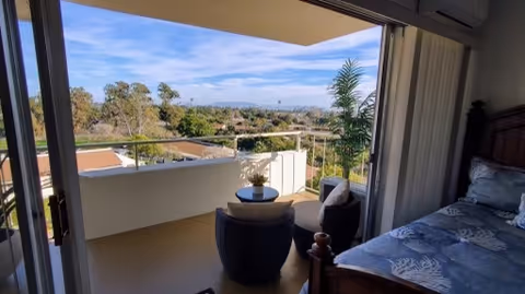 View from a bedroom looking out through sliding glass doors onto a balcony with a small round table and two chairs. The balcony overlooks a scenic outdoor area with trees and a clear blue sky.