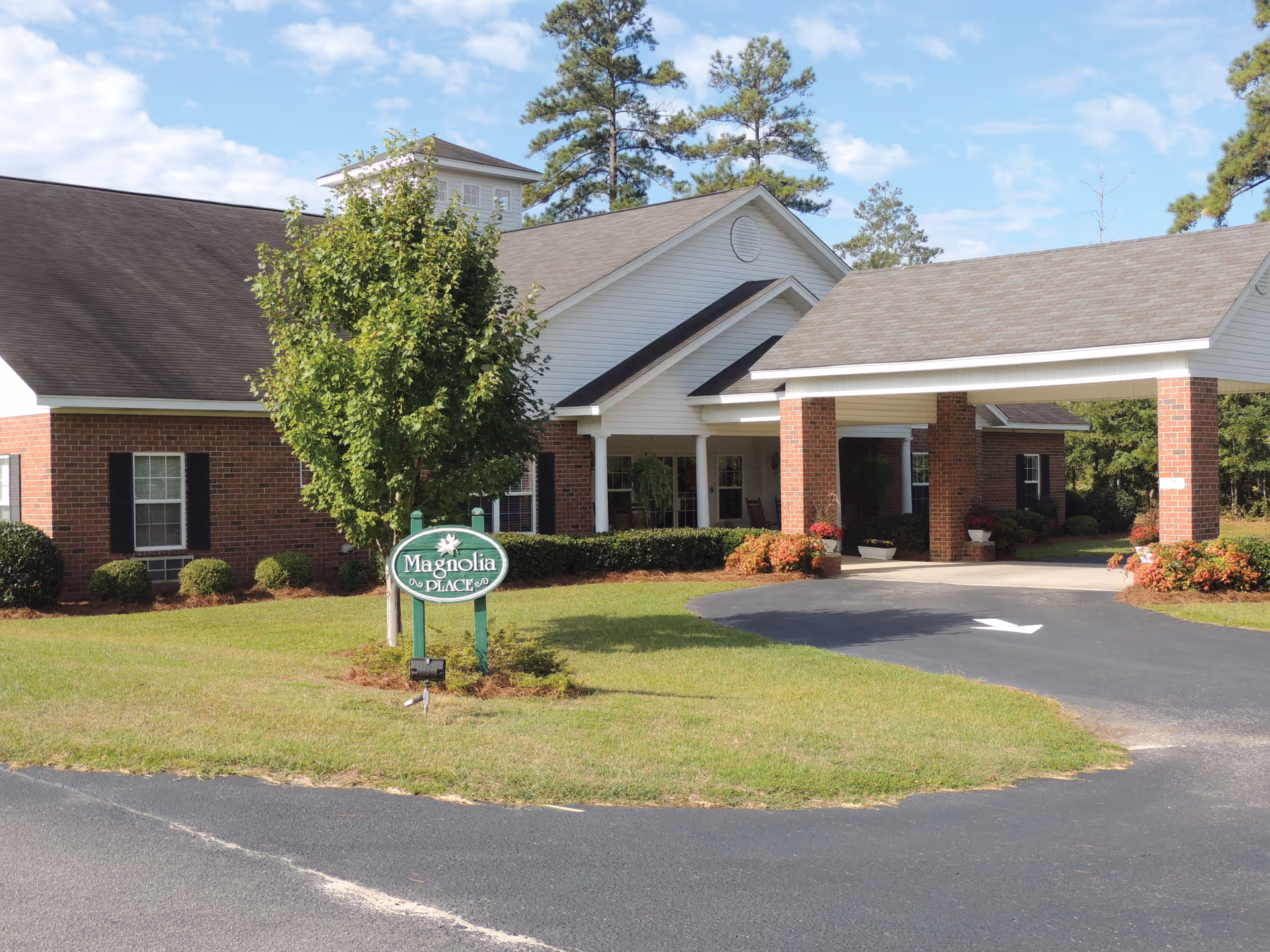 Exterior view of a single-story brick building with white trim and a covered entrance supported by brick columns. There is a green sign in front of the building that reads 'Magnolia Place'. The area is landscaped with grass, bushes, and a tree, and the sky is partly cloudy.