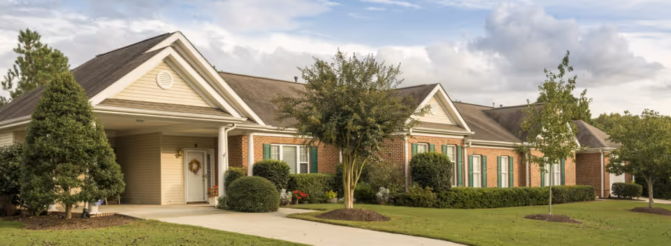 Exterior view of a single-story brick and siding building with a gabled roof, surrounded by well-maintained landscaping including trees, bushes, and a green lawn under a partly cloudy sky.