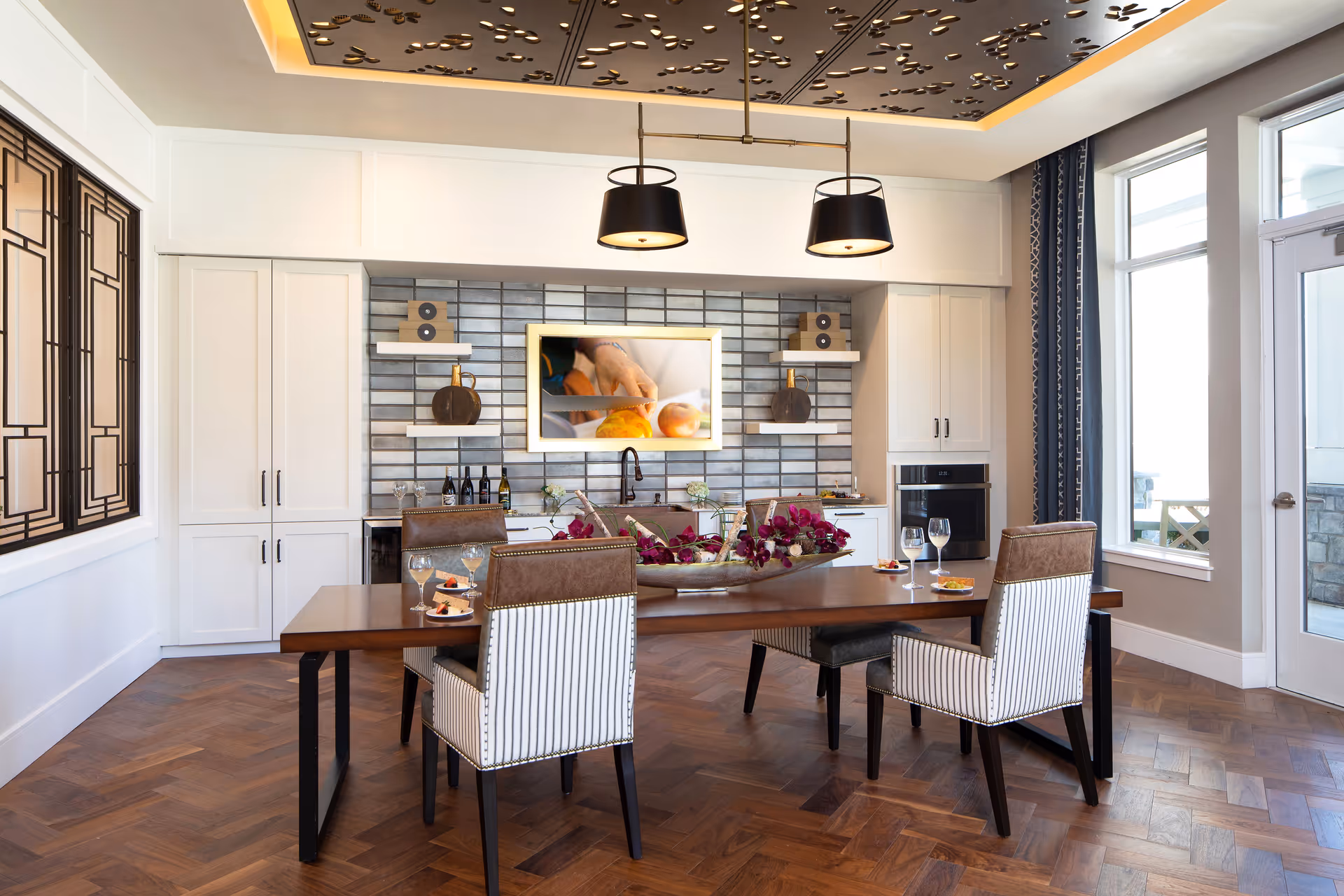 A modern dining room with a wooden table set for four, featuring striped and leather chairs. The room has a decorative ceiling panel with recessed lighting, a wall-mounted TV above a sink and countertop area with shelves holding decorative items, and large windows letting in natural light.