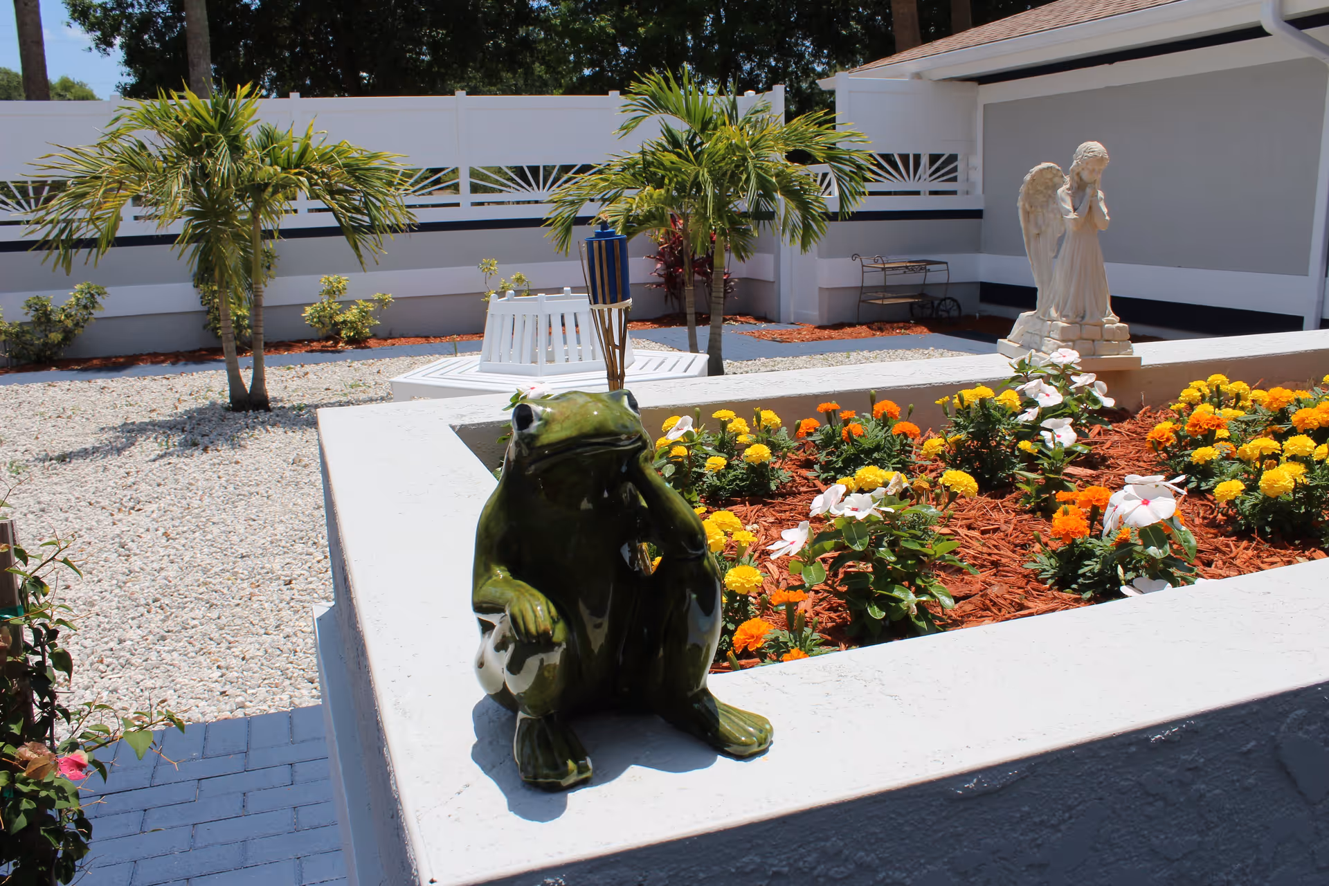 Outdoor garden area with a raised flower bed containing colorful flowers and a small green frog statue sitting on the edge. In the background, there are small palm trees, a white bench, a tiki torch, and a white angel statue. The area is surrounded by a white fence and has gravel ground cover.