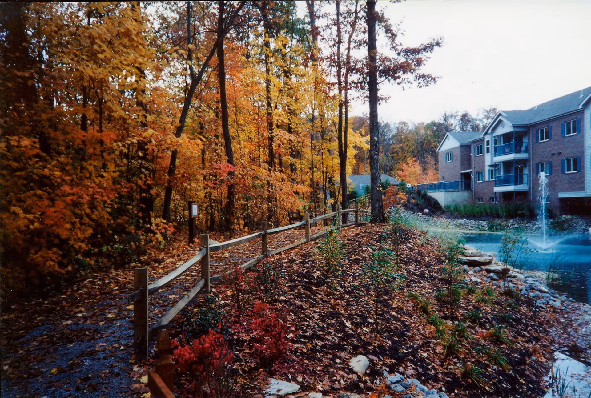 Wooded walkway in autumn beside a pond with a fountain and a multi-story brick senior living building.