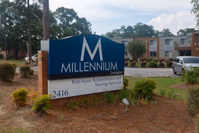 Outdoor view of the Millennium Post Acute Rehabilitation and Nursing Services sign with the building and parking lot in the background, surrounded by trees and landscaping.