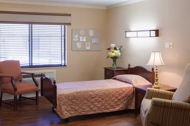 A cozy single-occupancy senior living bedroom with a twin bed, nightstands, chairs, a lamp, and a vase of flowers.