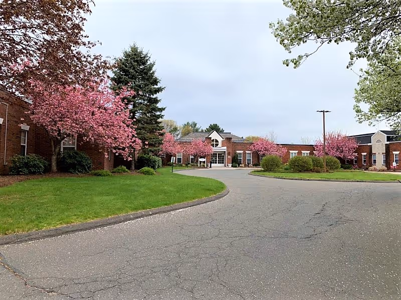 Exterior view of a single-story brick building with a curved driveway leading to the entrance. The building is surrounded by green grass, bushes, and trees with pink blossoms under a cloudy sky.