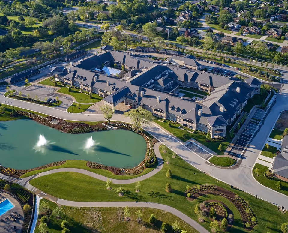 Aerial view of a large senior living facility named Flourish Collection at Rochester, featuring a multi-wing building surrounded by landscaped gardens, walking paths, a pond with two water fountains, and parking areas. The facility is located near a residential neighborhood with many trees and greenery.