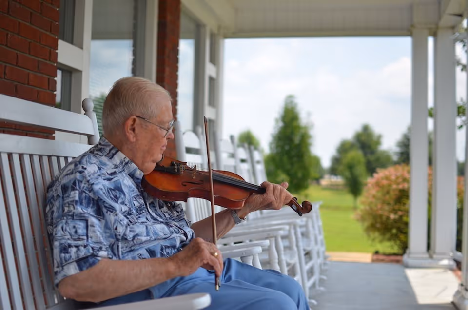 An elderly man sitting on a white rocking chair on a covered porch playing a violin. The porch has several white rocking chairs lined up, and there is a green lawn with trees and bushes in the background under a partly cloudy sky.