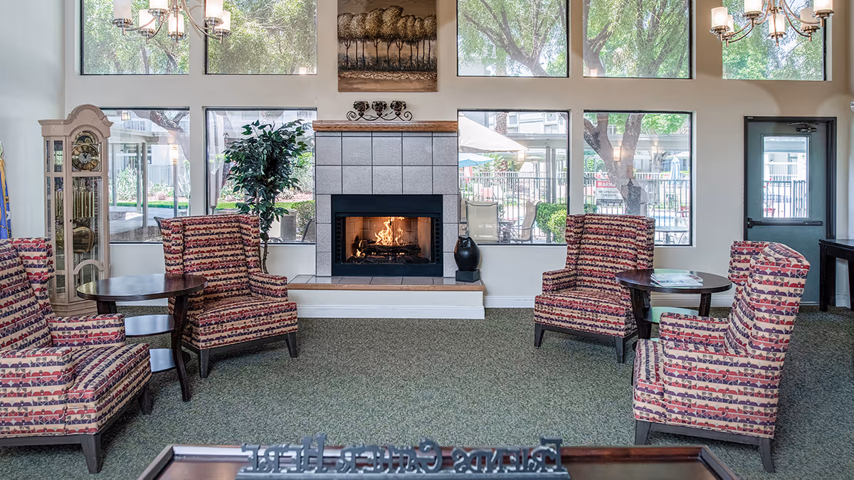 Sunny communal lounge with a central fireplace, patterned armchairs and side tables in front of large windows.