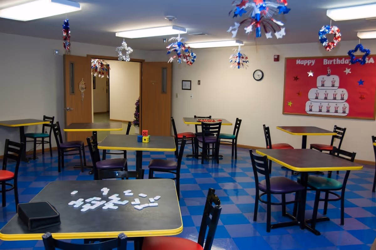 A community room with multiple tables and chairs arranged on a blue checkered floor. The tables have yellow edges and some have game pieces and dice on them. The room is decorated with red, white, and blue hanging decorations and a bulletin board with a birthday cake illustration and the words 'Happy Birthday'. There are double wooden doors leading to another room.