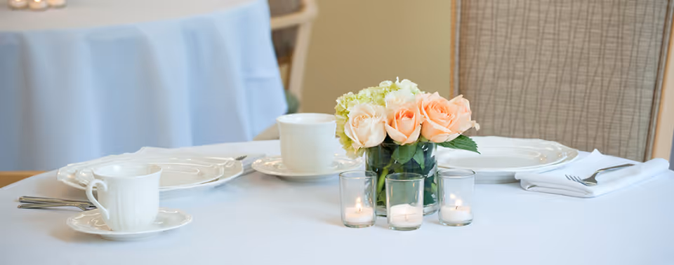 A dining table set with white plates, cups, and silverware on a white tablecloth. In the center, there is a small glass vase with peach and white roses and three small lit candles in glass holders. A cushioned chair is visible in the background.