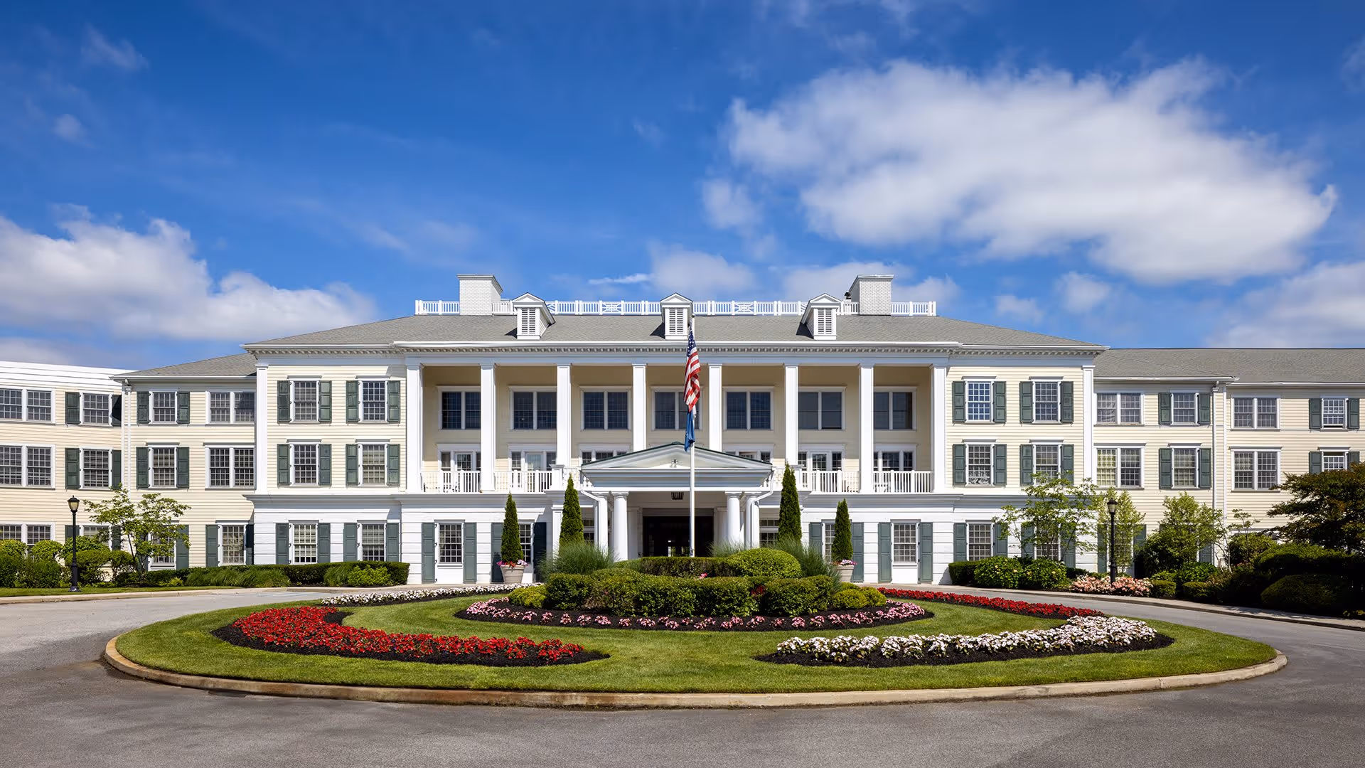 Front exterior view of a large, elegant senior living facility building with white siding, green shutters, and a central entrance with columns. The building is three stories tall with a manicured circular driveway and landscaped flower beds in front. An American flag is displayed on a flagpole in the center of the driveway. The sky is blue with some clouds.