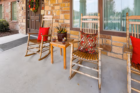 Front porch area of a building with two wooden rocking chairs, each with a decorative cushion, and a small wooden table between them holding a potted plant and two small decorative items. The building exterior features stone walls and a door with a fall-themed wreath.