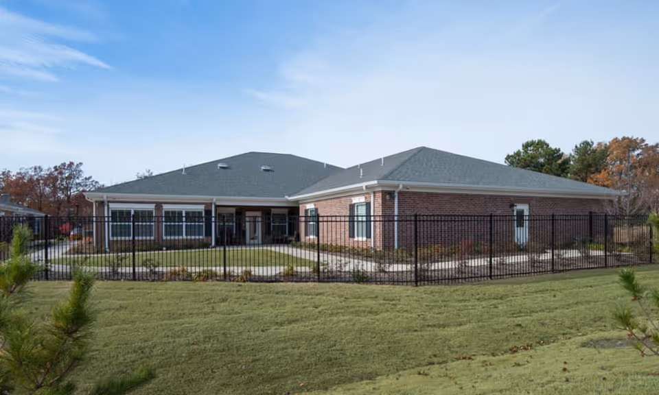 Exterior view of a single-story brick building with a gray roof, surrounded by a black metal fence and green lawn under a clear blue sky.